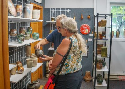 Two people examine a ceramic vase in a pottery shop filled with shelves of handmade pottery. The shop has gray walls, a window with a view of trees, and various pieces of pottery on display.
