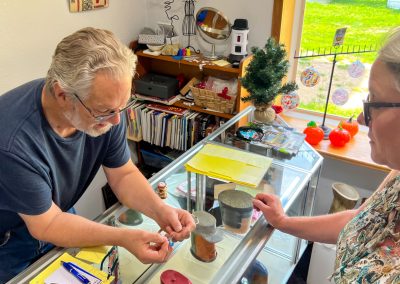 An older man behind a glass display case shows a small item to a woman in glasses at a shop. Shelves with books and decorations are visible, along with a window showing a green yard outside.
