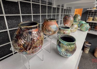 Colorful, handcrafted pottery vases with various patterns and glazes are displayed on white shelves against a dark, grid-patterned wall in a gallery or studio setting.