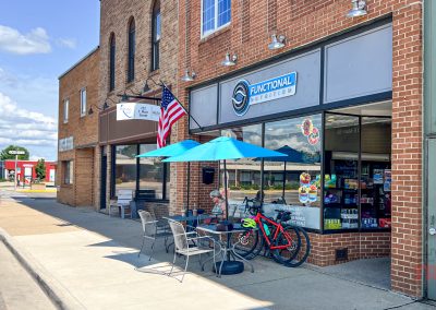 A brick storefront with an American flag, blue umbrellas, tables, chairs, and a red bicycle outside. The shop has a Functional Nutrition sign and large windows showing shelves of snacks inside.