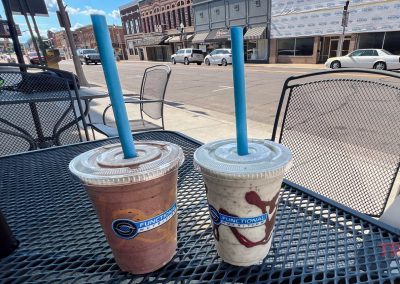 Two blended drinks with blue straws sit on a black metal outdoor table. The street and buildings are visible in the sunny background.