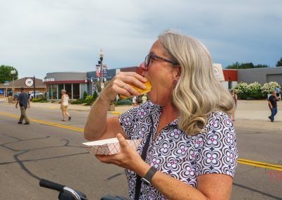 A woman with gray hair and glasses eats a hot dog from a paper tray while standing on a city street during an outdoor event. Other people and buildings are visible in the background.