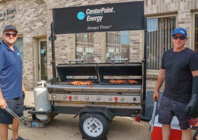 Two men holding grill tongs standing next to a grill trailer with hot dogs.