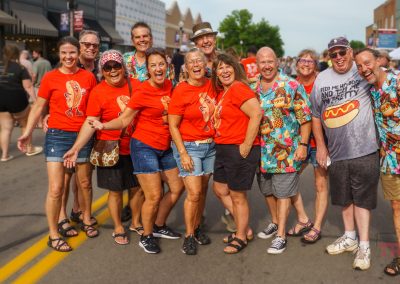 A group of people stand smiling in the street at an outdoor event. Most wear matching red t-shirts with hotdog graphics, while a few wear colorful shirts with hotdog patterns. Other festival-goers and tents are seen in the background.