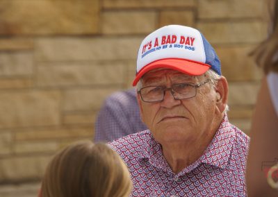 An older man wearing glasses and a cap that says ITS A BAD DAY TO BE A HOT DOG sits outdoors, looking serious. He is dressed in a red-patterned shirt, with blurred people and a stone wall in the background.