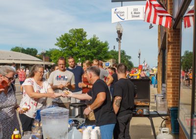 A group of people gather outside a brick building at a street fair, serving and receiving food from a table with drinks and condiments. American flags decorate the building, and a street sign reads “222 E. Main Street.”.