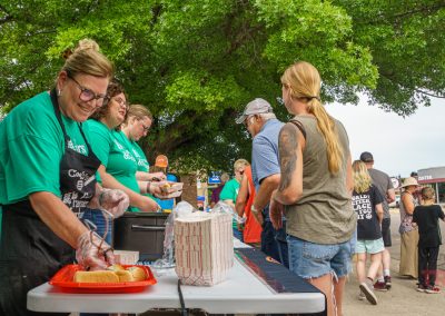 Volunteers in green shirts serve food from a table outdoors to people standing in line. A woman at the front of the table is smiling while preparing a plate with a hot dog. Trees and more people are visible in the background.