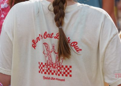 A person with a brown braid wears a white T-shirt with a red graphic of hot dogs and text that reads “Sun’s Out, Buns Out. Relish the moment.” The shirt has a red checkered pattern at the bottom.