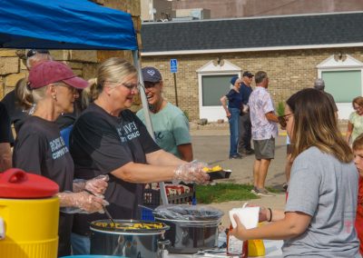 Volunteers serve food and drinks to people at an outdoor community event. One woman hands a plate to a guest, while others wait in line. The atmosphere is casual and friendly.