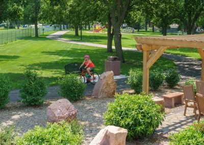 A woman wearing a helmet rides a bicycle on a paved park path lined with green grass and trees. In the foreground, there is a pergola, wooden chairs, and landscaped garden beds with large rocks.