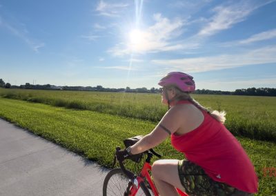 A person wearing a pink helmet and bright pink tank top rides a bicycle on a paved trail beside a grassy field under a clear, sunny sky.