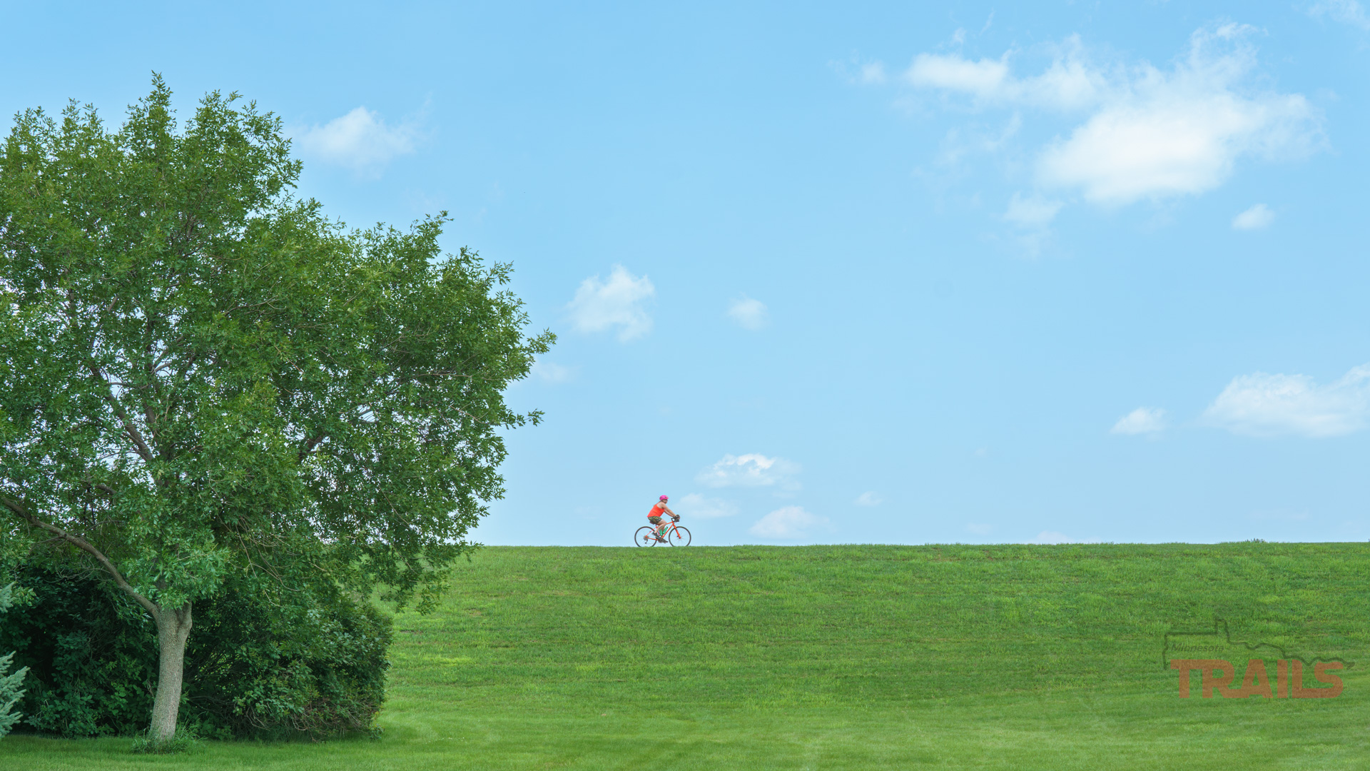 Luverne Loop Trail A person wearing a red shirt rides a bicycle along the top of a grassy hill under a bright blue sky with scattered clouds. A large leafy tree is on the left side of the image.