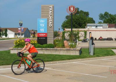 A person wearing a pink helmet and orange shirt rides a bicycle past the Trailhead sign for the Luverne Loop Trail in a small town, with buildings, greenery, and a clear sky in the background.