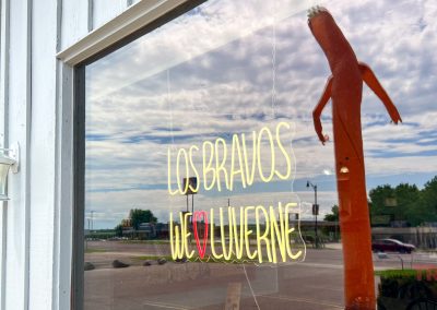 A window displays a neon sign reading LOS BRAVOS WE ♥ LUVERNE with reflections of the street, cars, and an orange inflatable tube man outside.