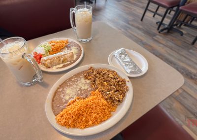 Two plates of Mexican food with rice, refried beans, and a taco on a table, along with two glasses of iced horchata, a wrapped tortilla, and empty chairs in a restaurant setting.