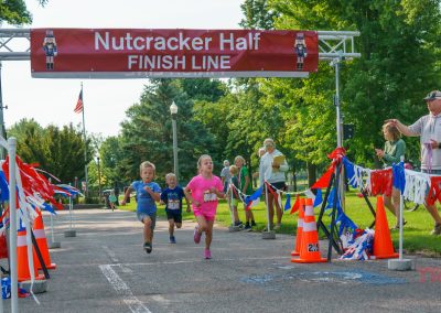 Three young children run toward the finish line of the Nutcracker Half marathon, cheered on by adults and volunteers. Colorful ribbons and cones decorate the path under a red finish line banner. Trees and a flag are in the background.