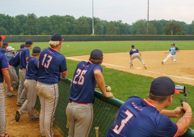 Baseball players in blue and gray uniforms watch intently from the dugout as a play unfolds on the field, with one player fielding the ball and another running toward a base on a sunny day.