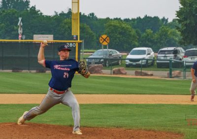 A baseball pitcher in a blue jersey prepares to throw a pitch on a baseball field, with a teammate in the infield and cars parked in the background near trees and a railroad crossing sign.