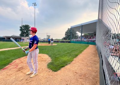 A young baseball player in a red helmet stands in the batter’s box, preparing to bat as spectators watch from bleachers and behind a chain-link fence on a grassy field under a partly cloudy sky.