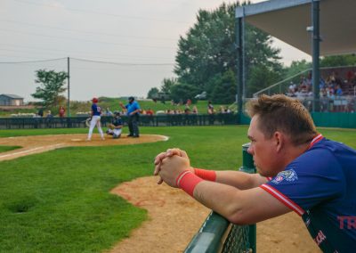 A baseball player in a blue jersey leans on a fence, watching a game in progress. In the background, a batter faces a pitcher as the umpire and catcher crouch behind home plate. Spectators sit in the stands.