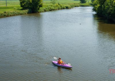 A person in an orange shirt paddles a purple kayak alone on a calm, tree-lined river under bright daylight, with green fields visible in the background.