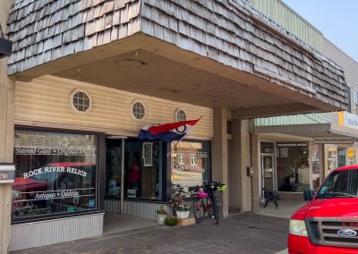 A storefront with a shingled overhang displays a sign reading Rock River Relics Antiques • Oddities. A red, white, and blue flag hangs outside. A red truck is parked in front and bicycles are visible near the entrance.