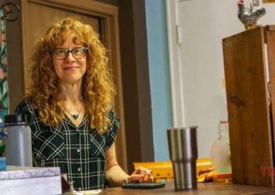 A woman with curly red hair and glasses stands behind a counter, smiling. She wears a plaid shirt and a necklace. On the counter are a tumbler, a water bottle, and a wooden box. The background is colorful and homey.