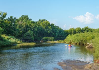 A person in a pink kayak paddles down a calm, tree-lined river on a sunny day with blue skies and scattered clouds.