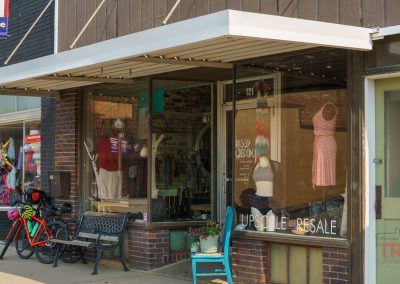 A storefront with large display windows showing mannequins dressed in clothing. A bench, bicycle, and colorful wreath stand are outside. A no parking sign and a Upscale Resale sign are visible in the scene.