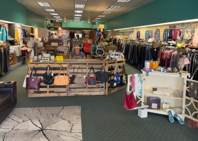 A boutique clothing store interior with racks of clothes, handbags displayed in front, shelves with accessories, and a person smiling behind the counter. The space is well-lit with teal and brick walls.