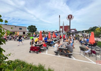 People sit at picnic tables with orange umbrellas in an outdoor patio area on a sunny day, enjoying food and drinks. Trees and buildings are visible in the background under a partly cloudy sky.