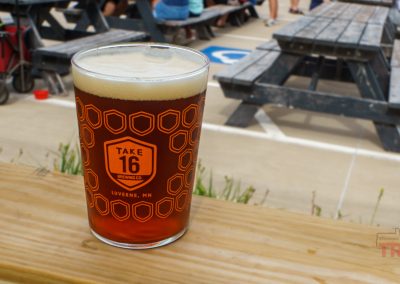 A pint glass of amber beer with a foamy head sits on a wooden railing outdoors. The glass has a honeycomb pattern and reads Take 16 Brewing Co. Luverne, MN. Picnic tables and people are in the background.