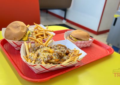A red tray on a yellow table holds two burgers, two servings of fries, and a serving of fries topped with melted cheese, all in paper baskets at a casual restaurant.