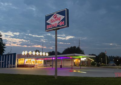 A retro-style drive-in restaurant with a large Tasty Drive-In sign and neon lights glows at dusk, with an empty parking lot and a cloudy evening sky in the background.