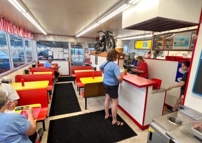 A retro-style diner with red and yellow booths, people dining, and two employees behind the counter. The decor includes vintage items like a bike and ice cream cone statue. The atmosphere appears casual and friendly.