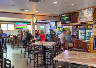 A lively bar with people sitting at tables and the bar counter, several TVs showing sports, and various drinks and decorations behind the bar. The atmosphere is casual and social.