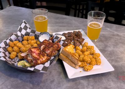 Two plates of food with tater tots, grilled meat, toast, and pickles are on a table next to two glasses of beer. Background shows chairs and partial view of people standing.