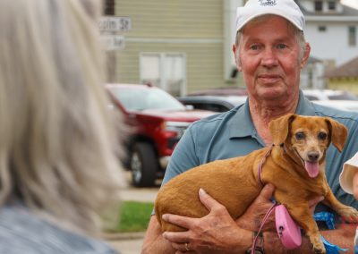 An older man stands outdoors holding a brown dachshund and a young child. A person with gray hair is in the foreground, and houses, cars, and street signs are visible in the background.
