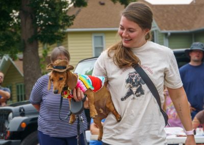 A woman smiles while holding a dachshund dressed in a colorful outfit and tiny cowboy hat at an outdoor event, with people and houses in the background.