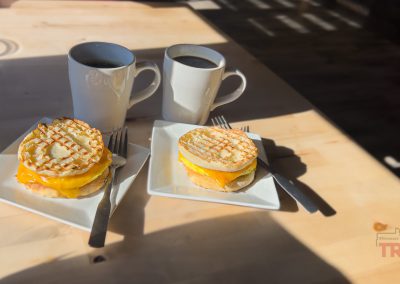 Two breakfast sandwiches with melted cheese on square plates and two white mugs of black coffee are placed on a light wooden table with sunlight and shadows. Each plate has a fork.