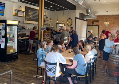 A group of people socializes inside a cozy café with wooden floors, a counter, and a refrigerator. Some sit around a central table, while others stand near the counter. Warm lighting and exposed brick walls create a welcoming atmosphere.