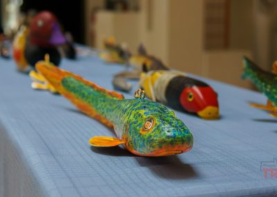 Colorful wooden fish carvings are displayed on a blue tablecloth, with the focus on a green and orange fish in the foreground. The background shows more fish carvings and appears softly blurred.