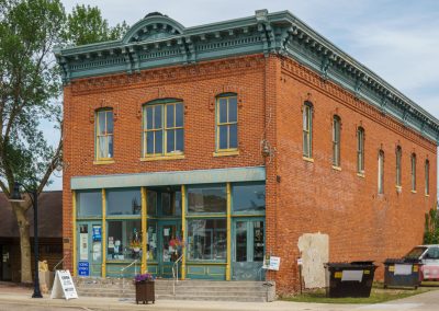 A two-story red brick building with large windows and decorative blue-green trim houses the New York Mills Regional Cultural Center. The entrance is flanked by flowers, and a tree stands to the left of the building on a sunny day.