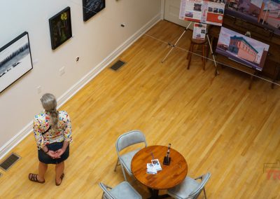 A person stands alone in an art gallery, viewing framed artwork on a white wall; nearby there is a round wooden table with chairs and informational materials, and more art displayed on easels in the corner.