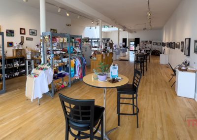 A bright art gallery and gift shop featuring wooden floors, art on white walls, shelves with crafts and clothing, and a small table with chairs and plants in the foreground.