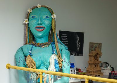 A large blue statue of a woman with braided hair, floral headband, and necklace stands in a room. Behind her is a black-and-white poster, a wooden model, and a white kettle on a countertop. Yellow railing is in front.