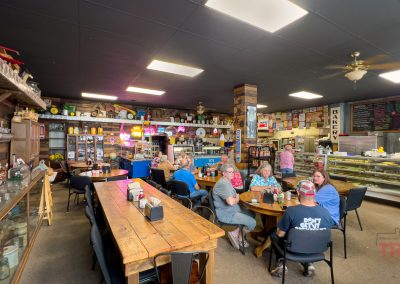 A group of people sit at wooden tables inside a cozy bakery café with shelves of decor, a display case of baked goods, and colorful menu boards on the walls. The atmosphere is casual and welcoming.