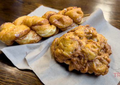 Two pastries rest on napkins atop a wooden table: a twisted, sugar-dusted doughnut and a chunky, golden-brown apple fritter.