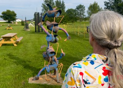 A person with gray hair in a ponytail, wearing a colorful, paint-splattered shirt, stands in front of a whimsical metal sculpture on a grassy field with picnic tables and other sculptures in the background.