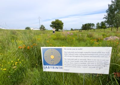 A sign in a grassy field explains a labyrinth walk, surrounded by wildflowers and tall grass. A few people walk in the background. The sky is partly cloudy and a wind turbine is visible in the distance.